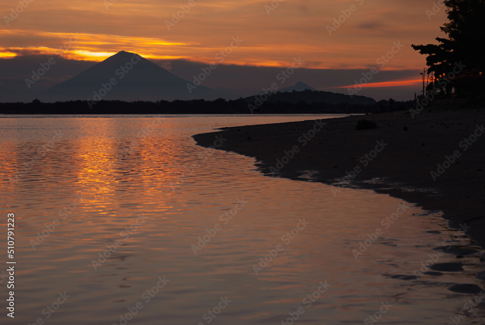 Bright majestic golden sunset on ocean with dark silhouette volcano, orange saturated reflection of sunbeams in water and black coastline. Dramatis indonesian landscape in evening, trip in asia.