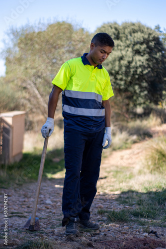 vertical portrait of an ethnic Latino municipal gardener with a tired expression looking down at the ground in uniform and holding a hoe in his hand, leaning on the ground.