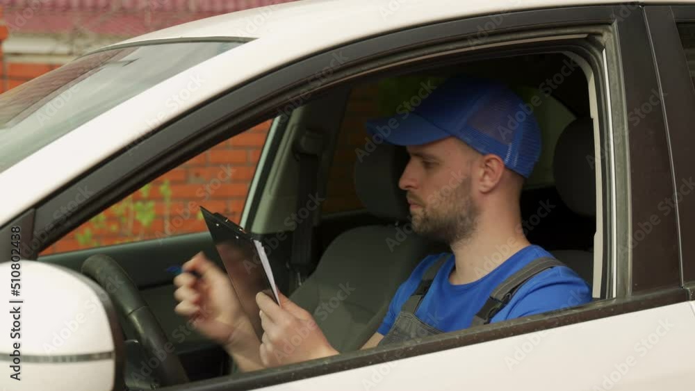 Master in cap writes data about car details from dashboard sitting on driver seat in white car. Person sits in auto and writes on clipboard with pen Master in cap writes data about car details from dashboard sitting on driver seat in white car. Person sits in auto and writes on clipboard with pen