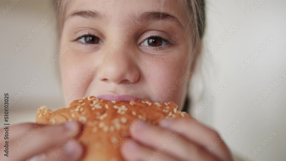 little kid girl eating a hamburger. unhealthy fast food meal proper lifestyle nutrition concept. child greedily with pleasure bites a big burger in the kitchen at home. kid eats fast food close-up little kid girl eating a hamburger. unhealthy fast food meal proper lifestyle nutrition concept. child greedily with pleasure bites a big burger in the kitchen at home. kid eats fast food close-up