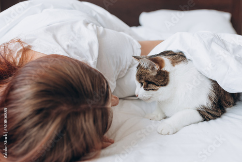 Obraz na płótnie Happy young woman with cat in bed at home. In cold weather, the pet warms up under a blanket. Pet friendly and grooming concept. Stray kitten sleep on bed. Cozy home background, morning bedtime. w salonie