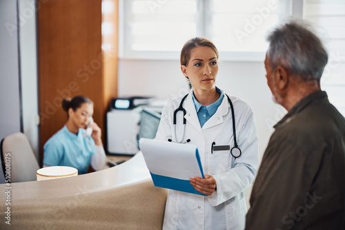 Female doctor talks to senior patient while going through his medical record at reception desk in hospital.