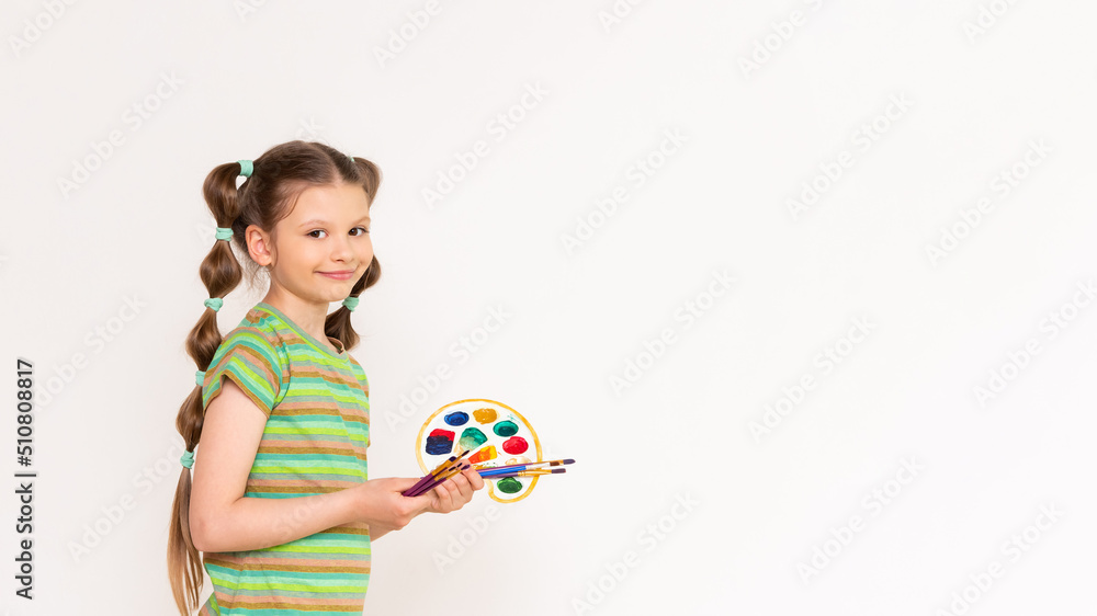 Drawing for schoolchildren. A little girl in a top with a palette of paints and brushes shows your advertisement on a white isolated background. Copy space. Drawing for schoolchildren. A little girl in a top with a palette of paints and brushes shows your advertisement on a white isolated background. Copy space.