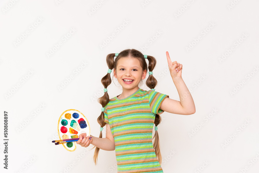Drawing for schoolchildren. A young artist with a palette and paint brushes points his finger up on a white isolated background.