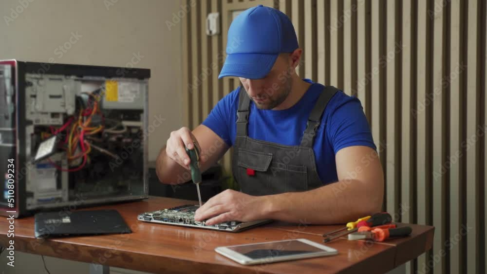Master in blue uniform installs internal details into tablet and closes cover with screwdriver sitting at wooden desk. Repair of tablet and computer electronics Master in blue uniform installs internal details into tablet and closes cover with screwdriver sitting at wooden desk. Repair of tablet and computer electronics
