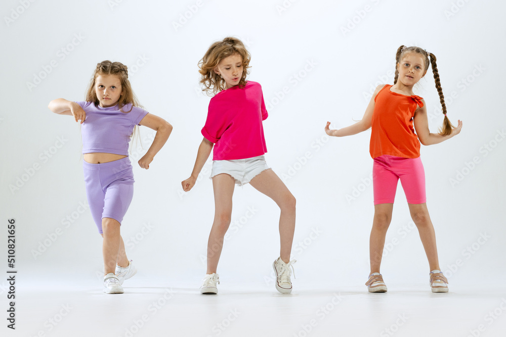 Fototapeta premium Three little girls, kids in bright colorful clothes dancing, posing isolated on white studio background. Concept of music, fashion, art, childhood, hobby