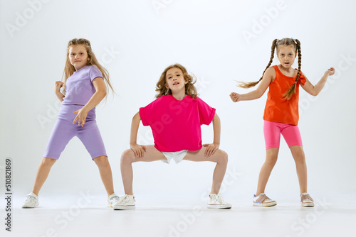 Three little girls, kids in bright colorful clothes dancing, posing isolated on white studio background. Concept of music, fashion, art, childhood, hobby
