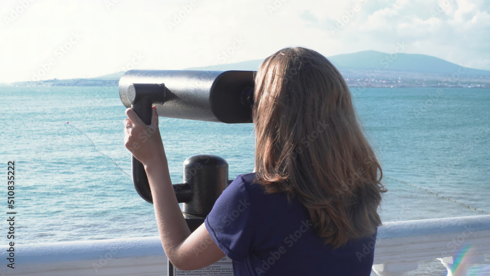 Beautiful woman looking through binoculars on background of sea and mountains. Media. Young woman views seascape with mountains in pair of binoculars on sunny day