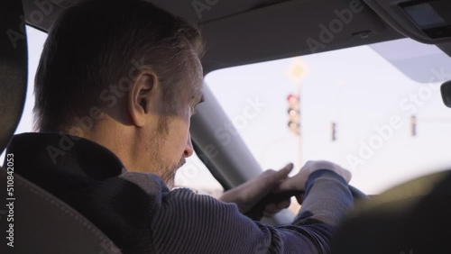 A middle-aged handsome Caucasian man waits impatiently at the red traffic light in a car, the light turns green and he drives on - rear closeup from the backseat, focus on the driver