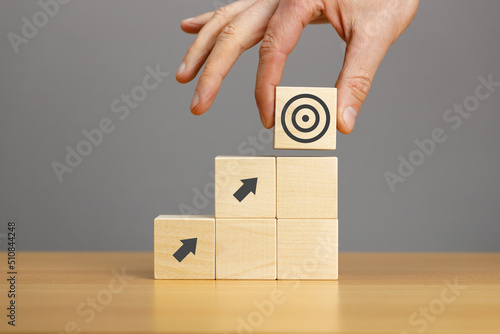 Hand putting wooden block pyramid with arrow and target icons on wooden table grey background. Business development strategy, advancement and goal concept.