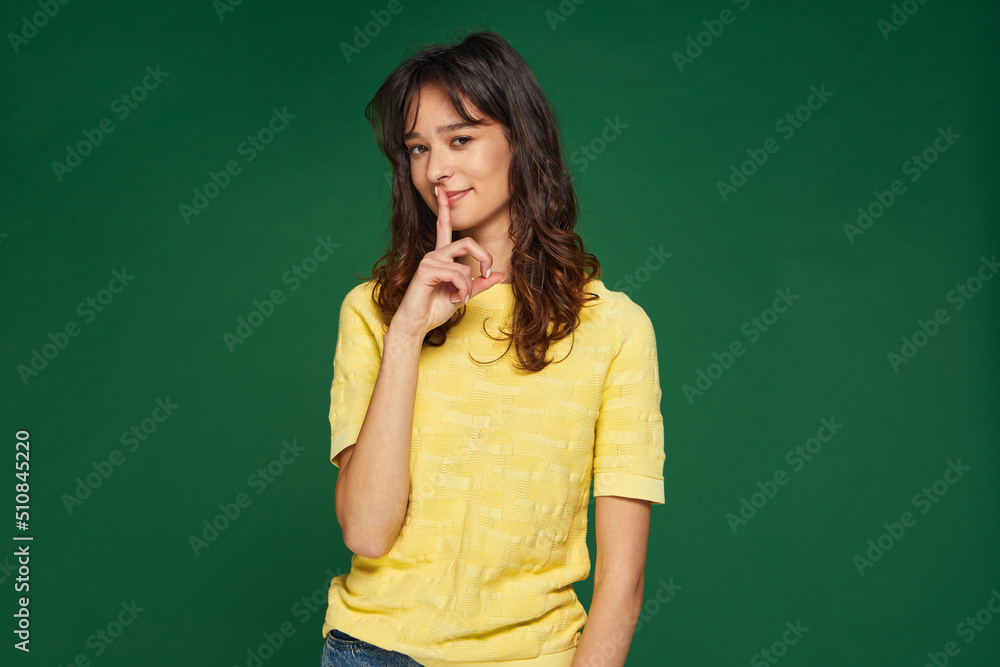 Smiling young girl showing silence gesture, ask to stay quiet, keep secret about surprise on green studio background Smiling young girl showing silence gesture, ask to stay quiet, keep secret about surprise on green studio background