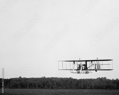 Fototapeta Wright Brothers biplane type B flying over a field with trees woods in the background