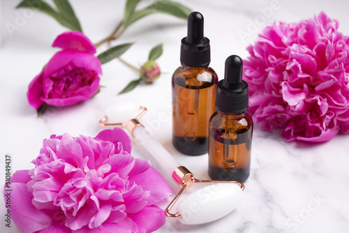 Glass bottles with oil, roller for face massage on marble background with peony flowers