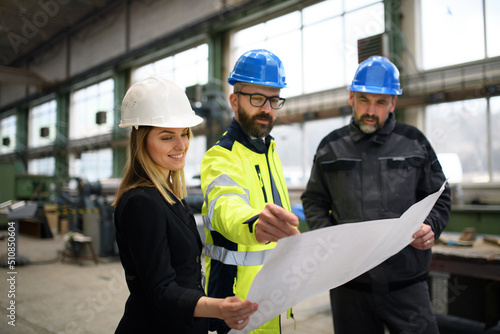 Manager supervisor, engineer and industrial worker in uniform discussing blueprints in large metal factory hall.