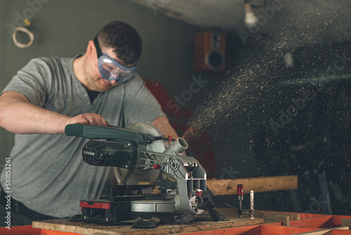 Strong male carpenter at work. The joiner holds the miter saw and a piece of plywood with his hand. Eyes are protected by special glasses. photo