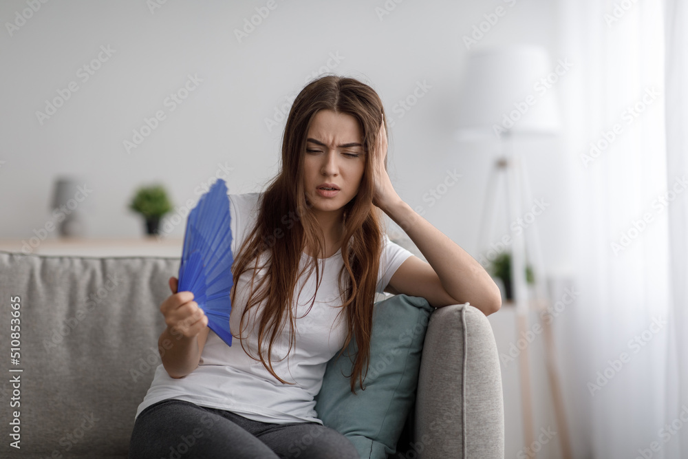 Sad young european woman suffer from hot and headache blowing fan at herself sitting on sofa in living room