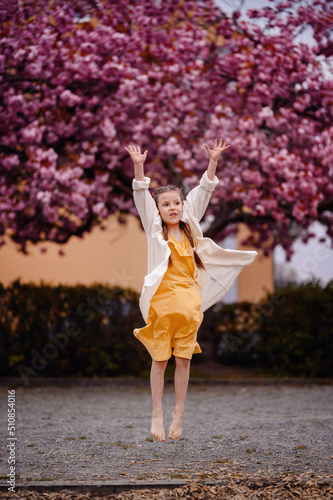Little preschooler girl in yellow dress, white cardigan with long hair stand before pink sakura tree