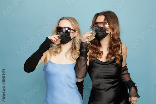 Fényképezés 2 Mädchen in eleganten Kleidern und mit Sonnenbrillen, trinken Champagner durch schwarze medizinische Masken hindurch