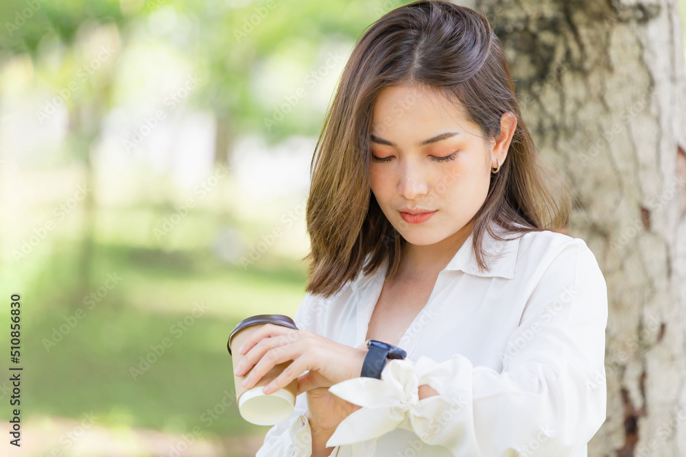 Young woman using smartwatch and holding drinks takeaway coffee while standing in garden under the tree Young woman using smartwatch and holding drinks takeaway coffee while standing in garden under the tree
