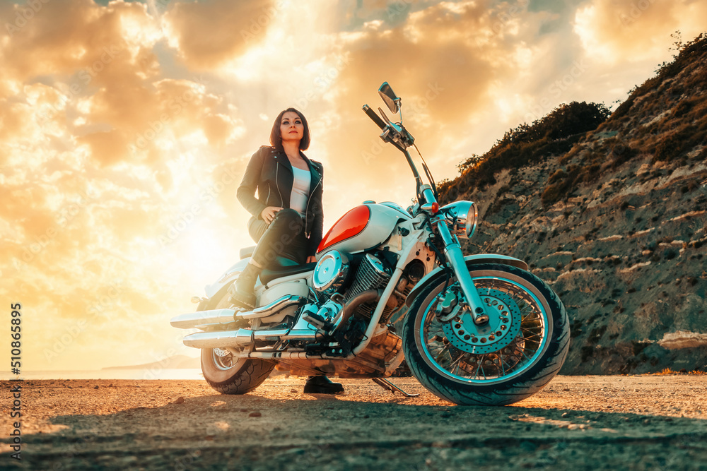 A beautiful confident adult woman in a leather clothes sitting on a vintage motorcycle. Bottom view. Epic sunset on the background. The concept of motorcycle travel and feminism