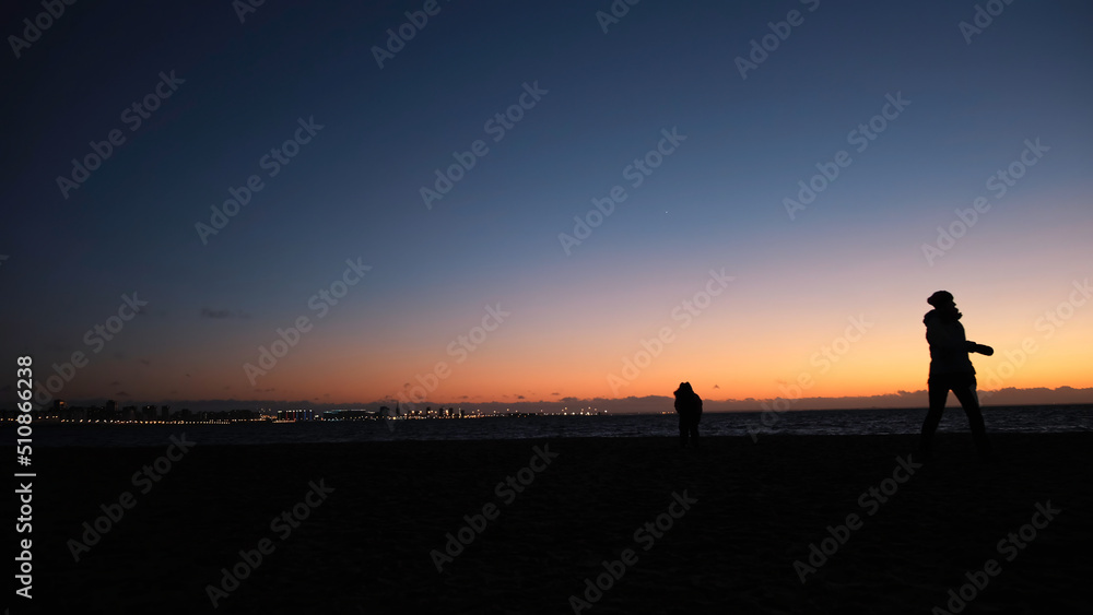 Close-up of mother's and child's silhouettes walking near the Gulf of Finland coast against the evening sky and city night lights. Concept. Beautiful sunset above the Gulf of Finland