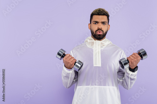 bearded african american sportsman exercising with heavy dumbbells isolated on purple.
