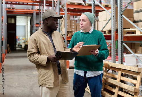 Female warehouse worker discussing list with delivery man while they standing in storage room