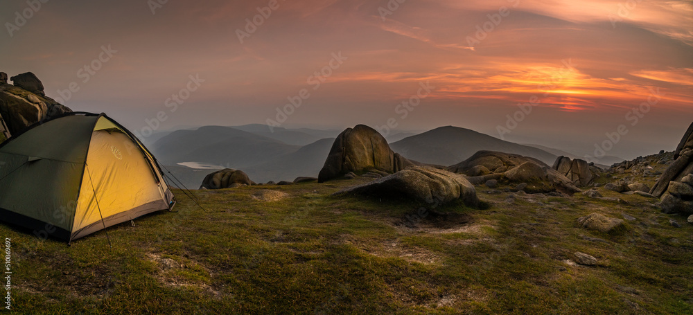 Glowing orange and green tent in the mountains under dramatic evening sky. Red, purple sunset and mountains in the background. Summer landscape. Panorama. Lake in the background.