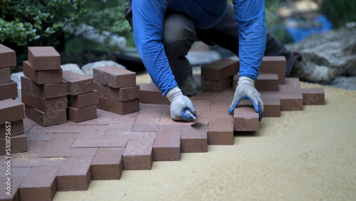 Using the butt of his trowel, a hardscaper taps the red brick pavers into place on a bed of sand in hardscaping landscaping patio project. photo
