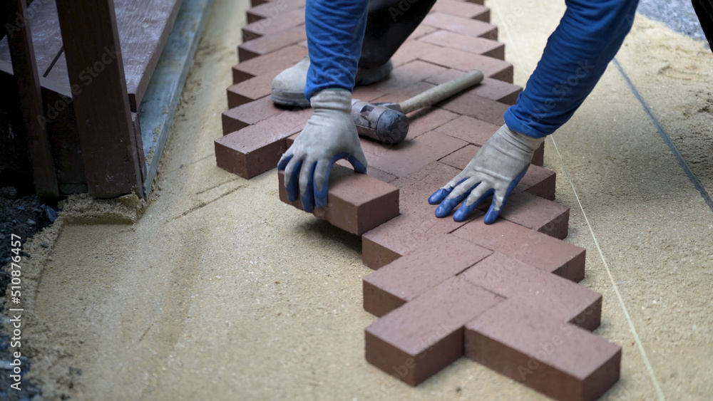 Man putting red brick pavers into place in a herringbone pattern on a bed of sand in hardscaping landscaping patio project. Man putting red brick pavers into place in a herringbone pattern on a bed of sand in hardscaping landscaping patio project.