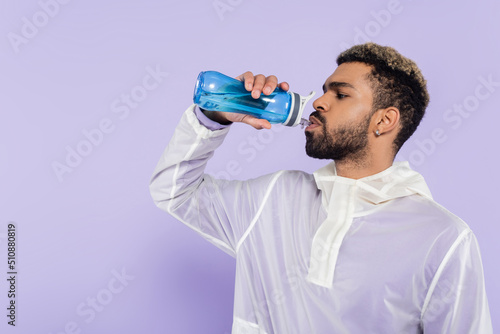 bearded african american sportsman holding sports bottle and drinking refreshing water on purple.