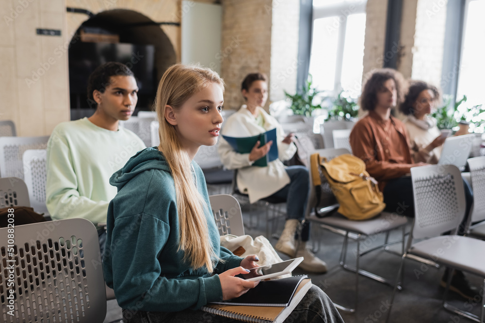 young woman sitting with notebooks and digital tablet near multiethnic students in classroom.