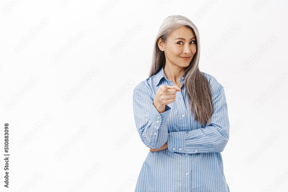 Smiling asian senior woman pointing finger at you, inviting clients, standing in casual clothes over white studio background Smiling asian senior woman pointing finger at you, inviting clients, standing in casual clothes over white studio background