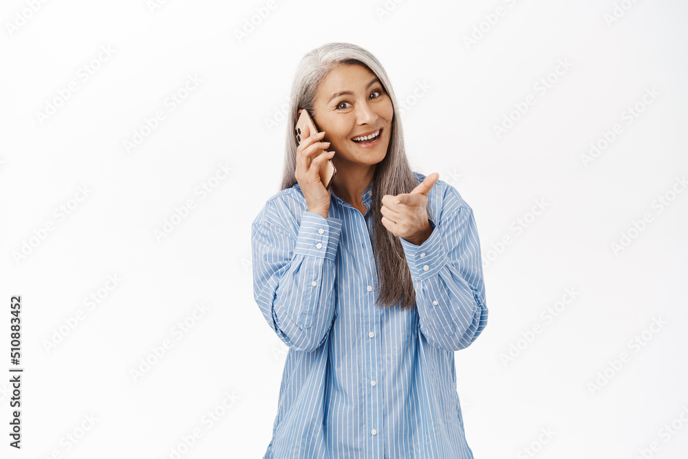 Happy asian old woman smiling, pointing at camera and talking on mobile phone, calling on smartphone, standing over white background Happy asian old woman smiling, pointing at camera and talking on mobile phone, calling on smartphone, standing over white background