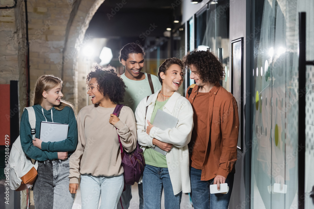 Fototapeta premium cheerful interracial students with backpacks smiling at each other in hallway of university.
