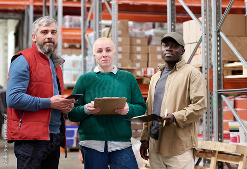 Portrait of group of multiethnic warehouse workers looking at camera while working with documents and tablet pc in storehouse