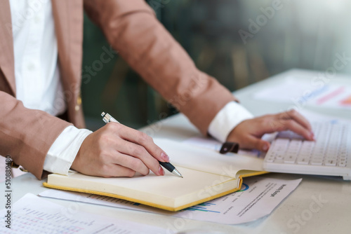 Close up image of a woman writing and taking note on notebook with laptop in office.