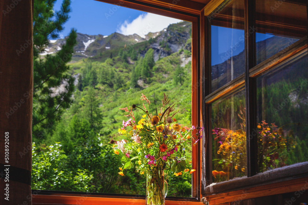 Fototapeta premium Blick aus einer Almhütte in die Landschaft auf die Berge. Im Fenster steht ein bunter Wiesenstrauß