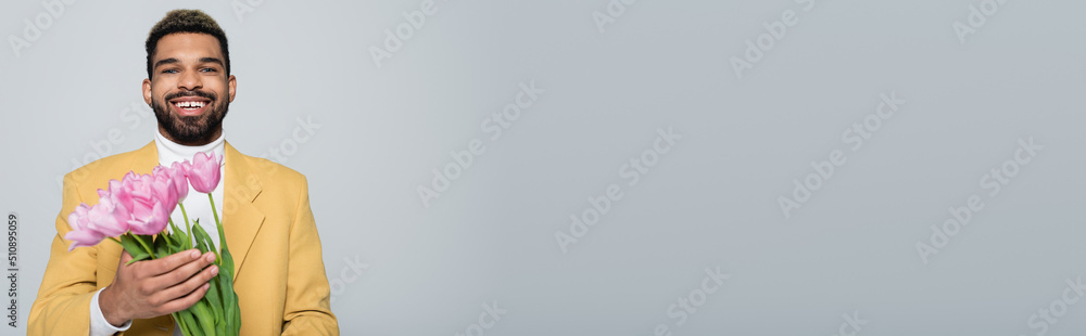 happy african american man in stylish outfit holding bouquet of pink tulips isolated on grey, banner. happy african american man in stylish outfit holding bouquet of pink tulips isolated on grey, banner.