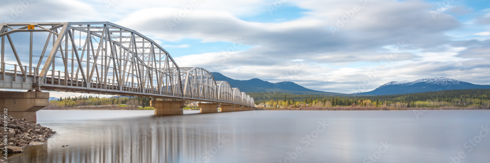 Large man made structure steel bridge spanning across Nisutlin Bay in township of Teslin flowing to the Yukon River in northern Canada during spring summer time with cloudy mountains background.	
