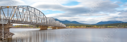 Large man made structure steel bridge spanning across Nisutlin Bay in township of Teslin flowing to the Yukon River in northern Canada during spring summer time with cloudy mountains background.   © Scalia Media