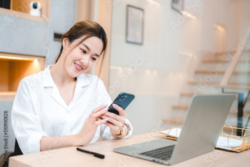 Smiling young Asian business woman and entrepreneur using mobile phone and scrolling and browsing through social media on taking break after working on laptop
