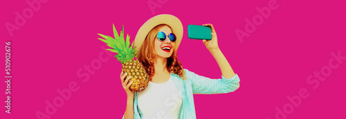 Summer portrait of happy smiling woman with pineapple taking selfie by smartphone wearing straw hat, sunglasses on pink background