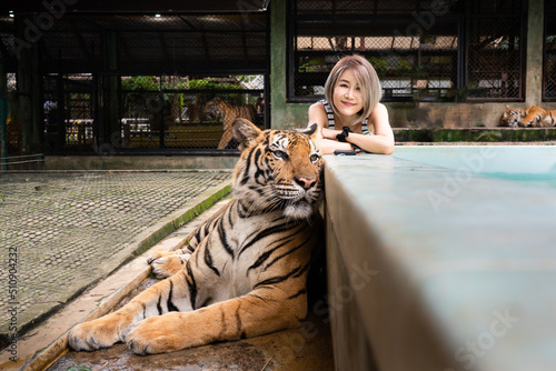 Selective focus of young short-haired Asian woman laying with arms crossed on the pool edge smiling at camera near orange-black striped adult tiger laying on ground in front of her at Tiger kingdom.