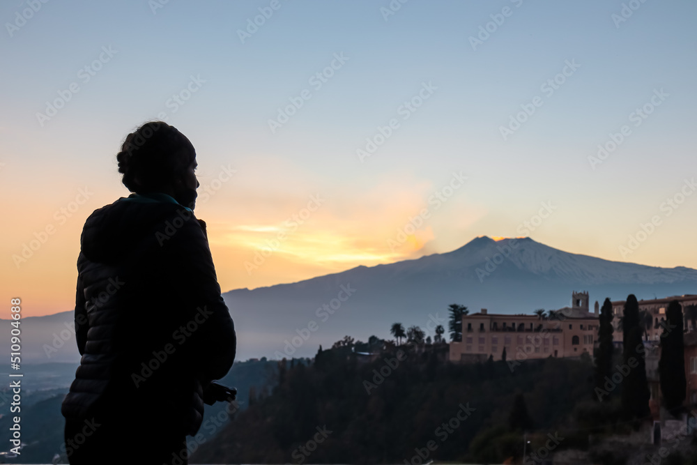 Fototapeta premium Tourist woman at a decorative railing with scenic view on snow capped Mount Etna volcano during sunset from public garden Parco Duca di Cesaro to Giardini Naxos in Taormina, Sicily, Italy, Europe, EU