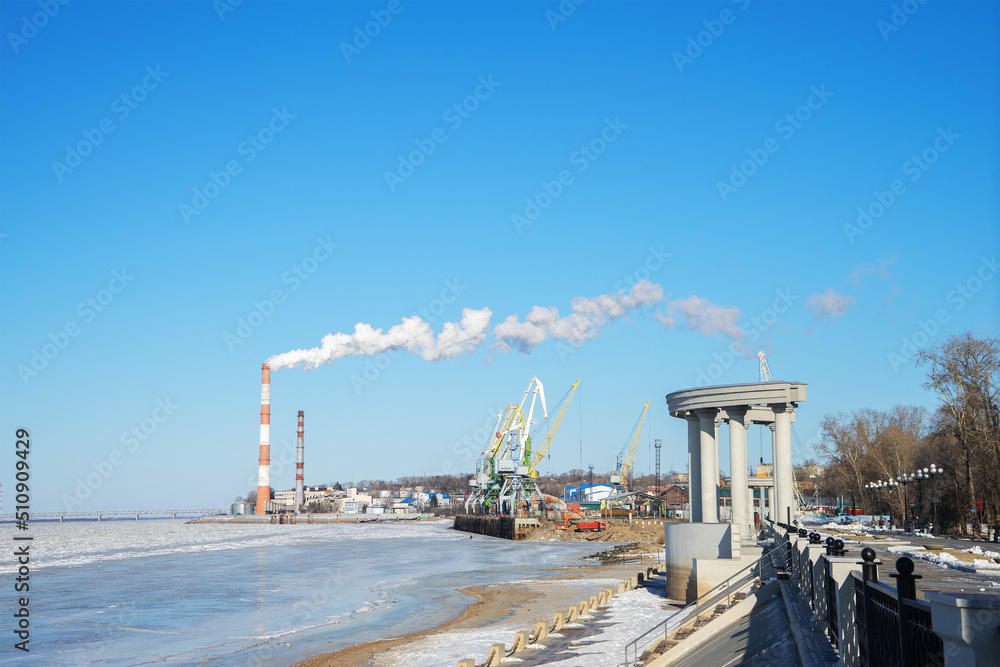Obraz premium Embankment of Amur River, covered with ice on clear winter day, Khabarovsk. White smoke from chimneys of factories on riverbank, port cranes. Сoncept of ecology