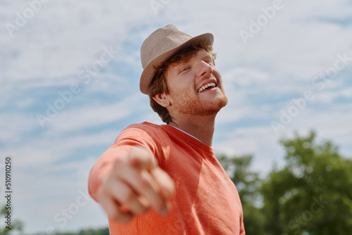 Happy young redhead man in hat pointing camera and smiling while standing outdoors © gstockstudio