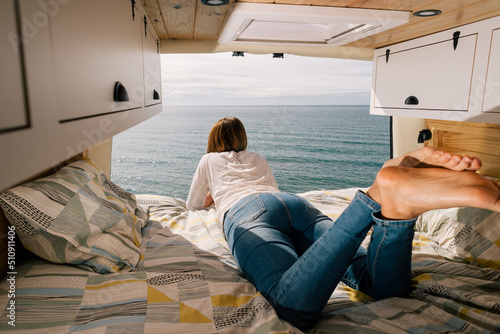 Woman lying on the bed of a camper van with the doors open looking at the seascape.