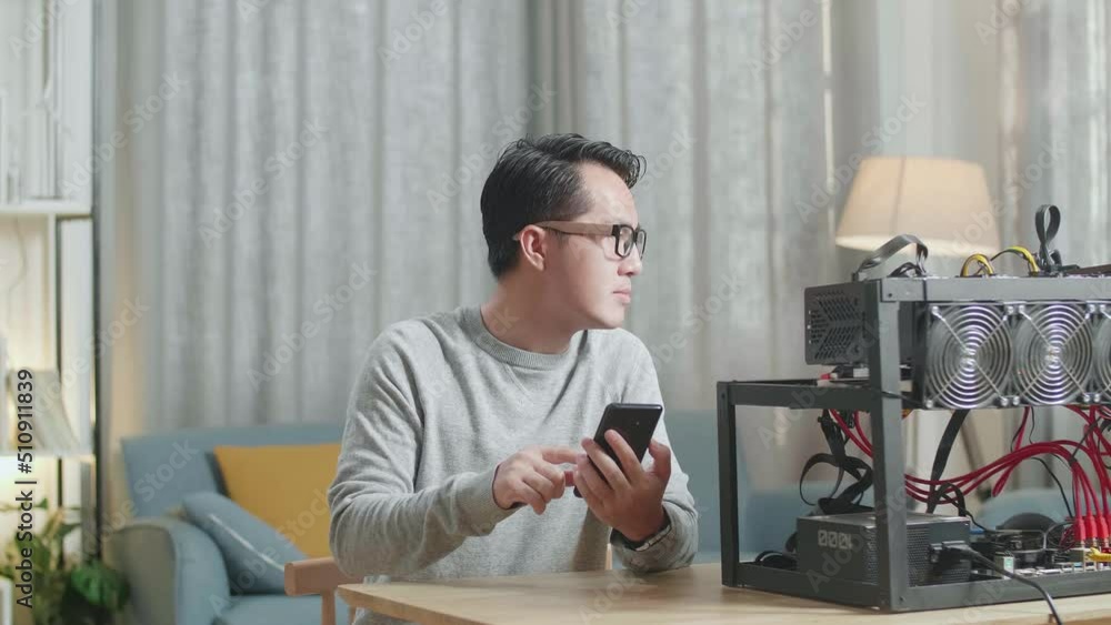 Asian Man Wearing Glasses Smiling And Shaking His Head While Using Smartphone With Mining Rig Mining Cryptocurrency
Asian Man Wearing Glasses Smiling And Shaking His Head While Using Smartphone With Mining Rig Mining Cryptocurrency