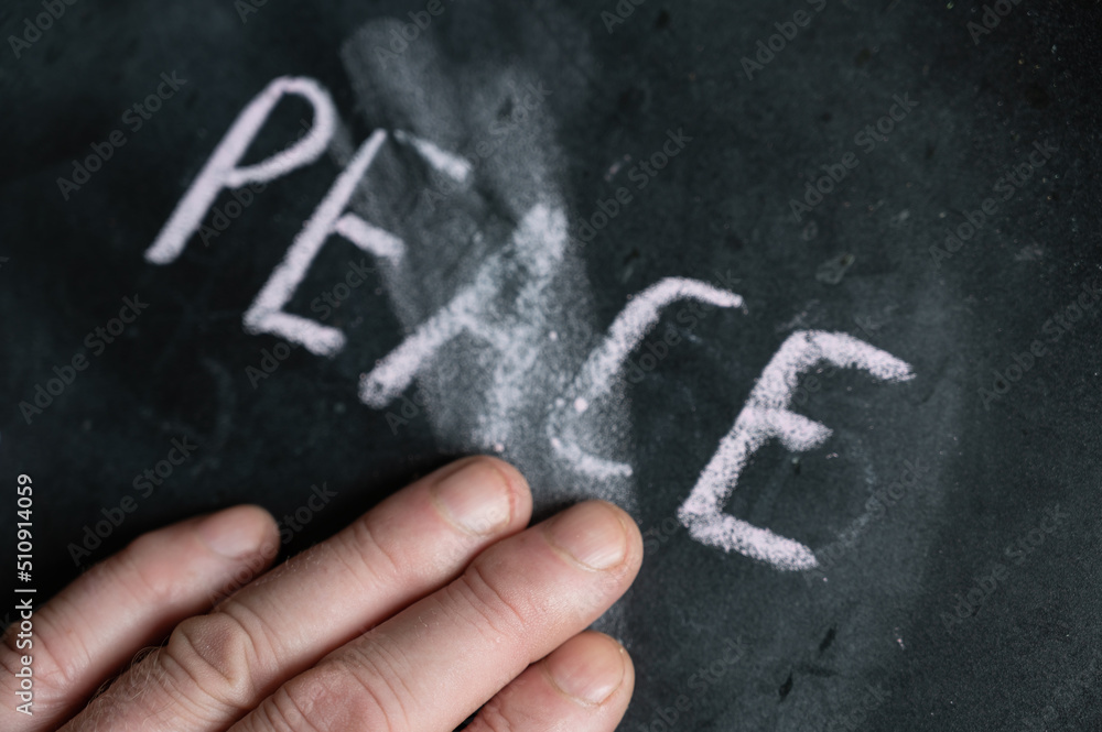 The semi-erased word PEACE on the black chalkboard. An adult man's left hand removes the handwritten word with his fingers. Fuzzy letters on black surface.